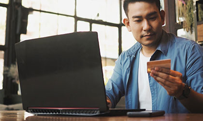 Smart young Asian man using credit card on laptop while drinking coffee at a desk
