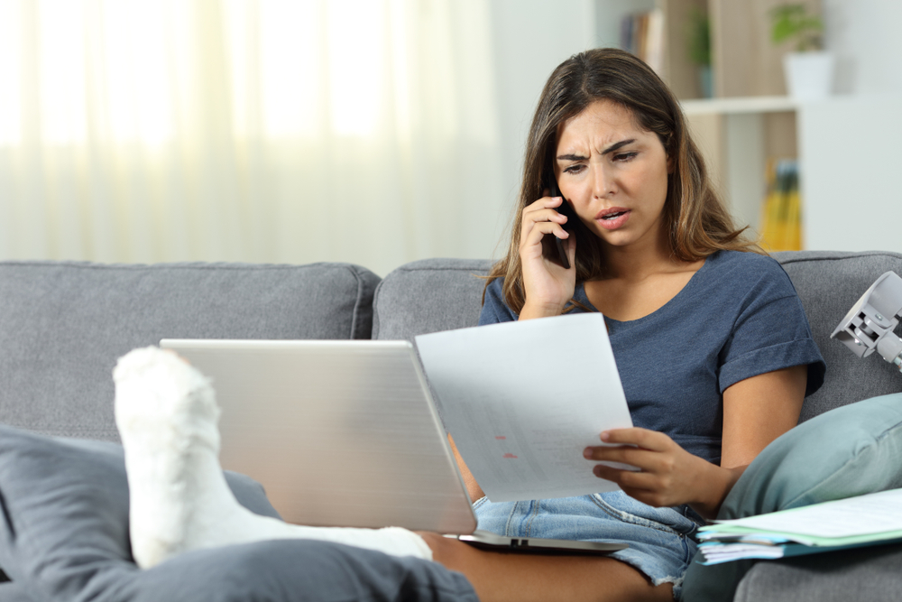 Stressed woman on phone looking at paperwork and laptop with leg in cast elevated.