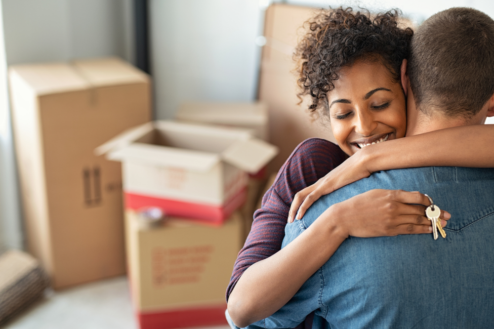 Young black woman embracing man and holding home keys with boxes in the background.