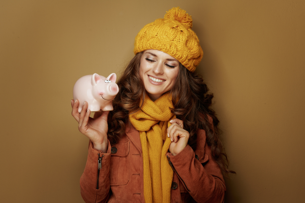 Young women in yellow knit hat and scarf holding and smiling at piggy bank