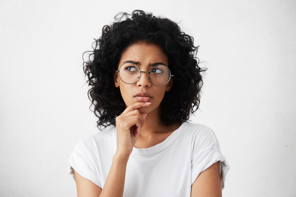 Young women with dark, curly hair and glasses touching her chin with a confused expression.