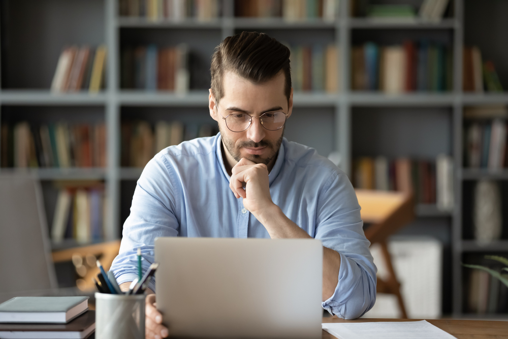 Caucasian man wearing glasses thoughtfully looks at computer screen.