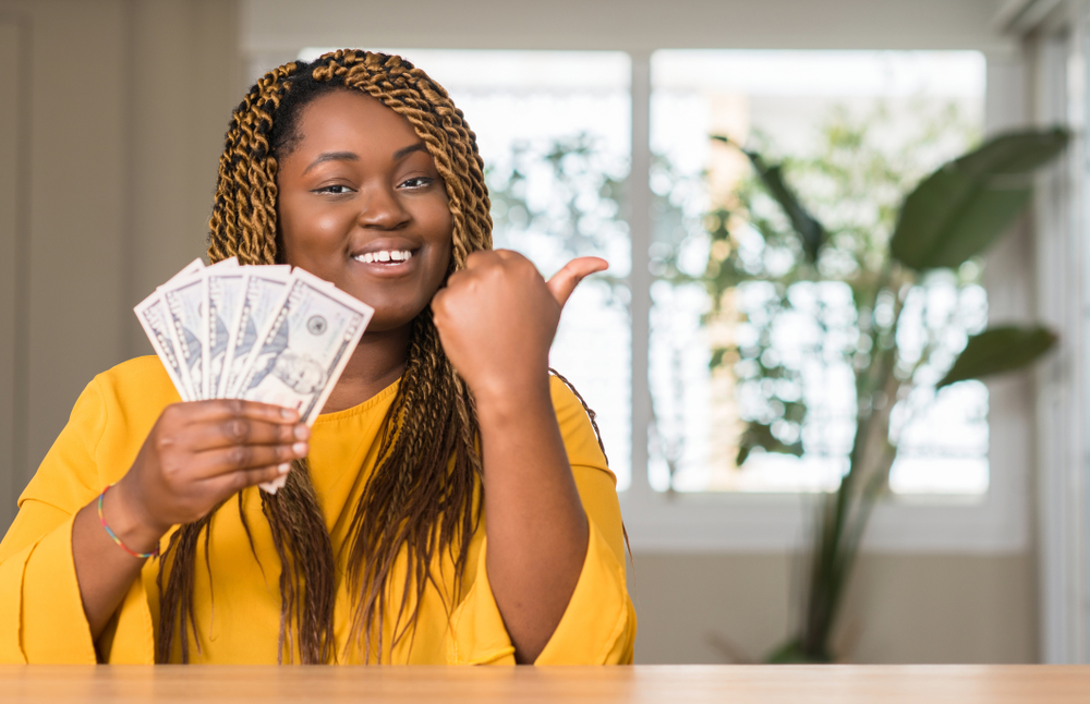 Young smiling Black woman holding cash with a thumbs up.