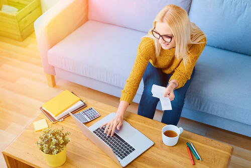 Happy young woman paying bills on laptop sitting on sofa