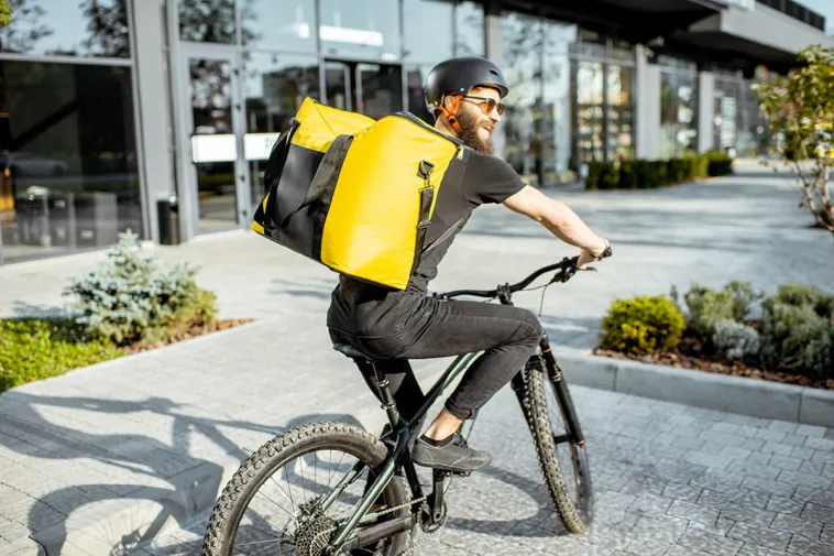 A smiling bicycle delivery worker wearing a yellow insulated bag rides through an urban area, representing gig economy side hustles as a way to earn extra cash.
