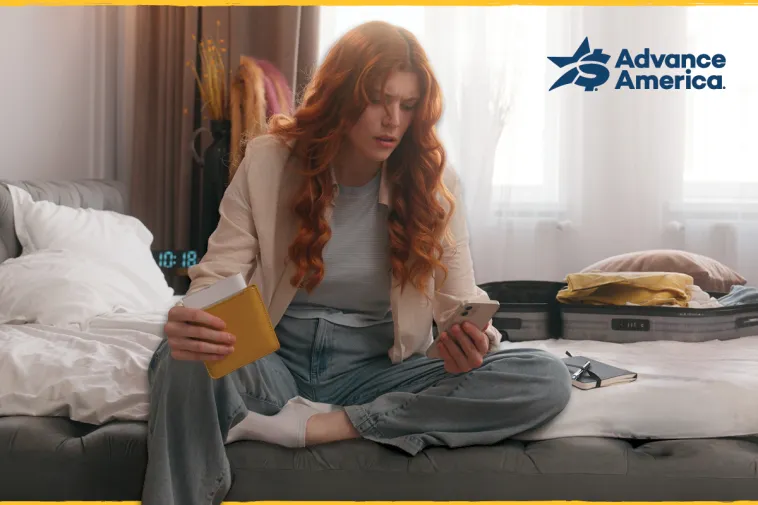 A woman with long red hair sits cross-legged on a bed, holding a yellow wallet and looking at her smartphone. An open suitcase with folded clothes sits beside her, and a notebook rests nearby. The Advance America logo appears in the upper right corner.