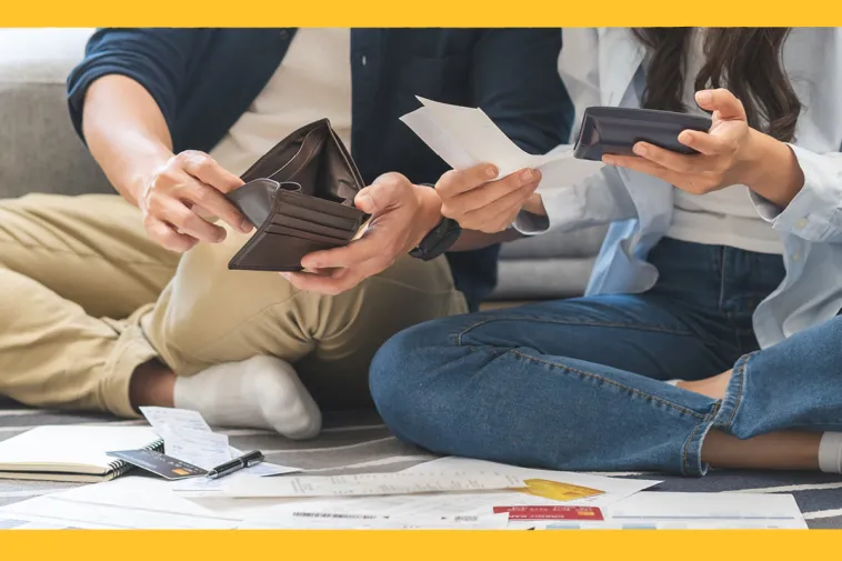Two people sitting on the floor surrounded by bills, receipts, and credit cards, reviewing their finances together — one holding an open wallet and a receipt, the other holding a phone.