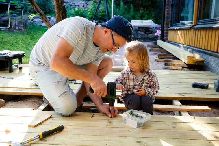 A young dad and daughter sit on the floorboards of a home deck as he demonstrates how to screw in a nail.