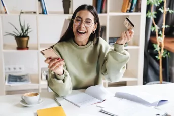 Cheerful student with long hair and glasses holds up her phone and credit card and smiles.