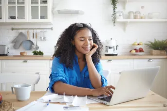 Happy Black woman sits at kitchen table looking at laptop.