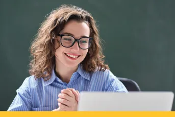 A smiling woman with curly hair and glasses sits at a desk with her hands clasped, looking confidently at a laptop screen.