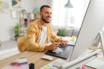 A smiling young man works at a home desk using a drawing tablet and desktop computer. Color swatches, sticky notes, and design materials are spread across the desk, suggesting he is a freelance creative professional working from home.