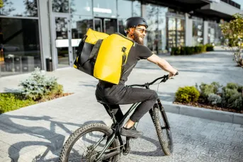 A smiling bicycle delivery worker wearing a yellow insulated bag rides through an urban area, representing gig economy side hustles as a way to earn extra cash.