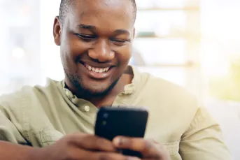 A young man smiles broadly while looking at his smartphone, sitting in a bright, softly lit space — conveying ease and confidence after applying for a same-day loan online.