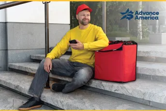 A food delivery worker in a yellow sweatshirt and red cap sits on outdoor steps holding a smartphone, with a red insulated delivery bag beside him. The Advance America logo appears in the upper right corner.