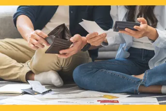 Two people sitting on the floor surrounded by bills, receipts, and credit cards, reviewing their finances together — one holding an open wallet and a receipt, the other holding a phone.